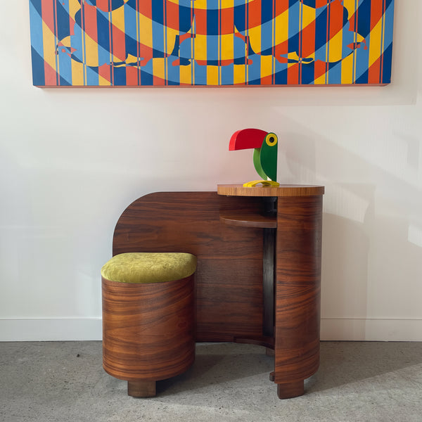 Art Deco Walnut vanity with a green and red parrot figurine, next to a round stool with a green cushion, against a white wall with colorful abstract art.